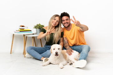 Young caucasian couple with dog staying at home smiling and showing victory sign