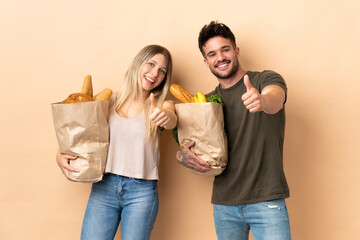 Couple holding grocery shopping bags over isolated background giving a thumbs up gesture because something good has happened