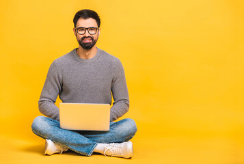 Portrait of a happy young bearded man in casual holding laptop computer while sitting on a floor isolated over yellow background.