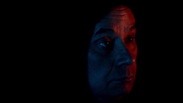 elderly man in a dark room with neon lights in a black hood sits, watches TV, grins, head close-up, concept mood, modern technologies, emotions, addiction, adrenaline