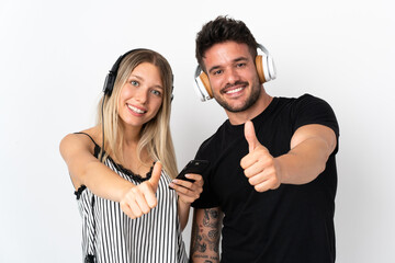 Young caucasian couple on white background listening music and with thumb up