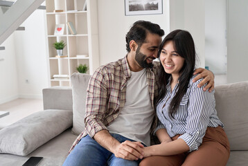 Happy affectionate indian couple bonding, hugging and laughing sitting on couch at home. Smiling young husband and wife embracing, having fun together, enjoying talking relaxing on sofa in apartment.