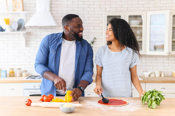 Overjoyed black couple standing next to kitchen counter, spending weekend together at home, looking at each other, cooking, girl responsible for spreading sauce on pizza, guy chopping veggies peppers