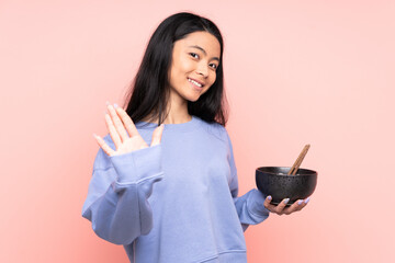 Teenager Asian woman isolated on beige background saluting with hand with happy expression while holding a bowl of noodles with chopsticks