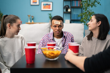 In the Living Room Diverse Group of Fun Loving Friends Playing Who am I game With Sticky Papers Attached to Foreheads. Guys and Girls Trying to Guess What is Written on Sticky Notes