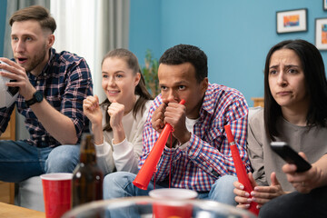 A young African-American male student keeps his fingers crossed for his football team. A group of friends of different nationalities watch the game while sitting on the living room couch at home.