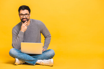 Portrait of a happy young bearded man in casual holding laptop computer while sitting on a floor isolated over yellow background.