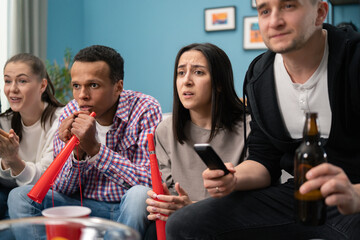 A young hindu female student keeps his fingers crossed for his football team. A group of friends of different nationalities watch the game while sitting on the living room couch at home.