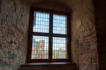 Fototapeta na wymiar Autumn day light going through vintage stained-glass window in very thick wall niche in the dark medieval castle. Ruins of medieval castle behind the window.