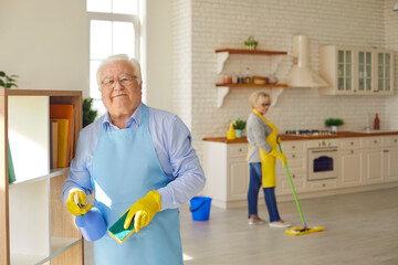 Senior man wipes the shelves holding a washcloth and spray with detergent while his wife on the background washes the floor. Concept of neatness, tidiness and cleanliness. Blurred background.