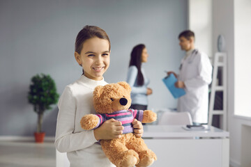 Close up of smiling healthy little girl hugging soft toy bear in doctor's office looking at camera....