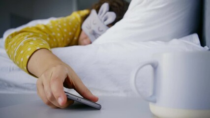 Sleepy young woman in grey eye mask with funny bunny face and color pajamas seeks phone to turn off alarm clock in bedroom in early morning - Powered by Adobe
