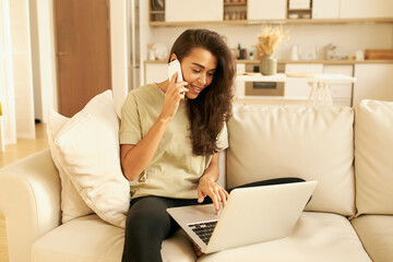 Stylish young Arabic woman entrepreneur multitasking while working distantly from home, sitting on sofa with laptop, checking something on website and answering phone call, speaking to customer
