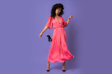 Elegant black woman in pink  party dress having fun in studio over  purple background.  Wearing heels. Full lenght.