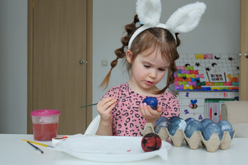 An adorable little girl in a dress and bunny ears is concentrating on Easter eggs at home. Easter preparation concept. Happy easter