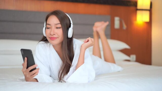 Happy pretty Asian woman watching phone and listen to music while lying on the hotel bed wearing a white dressing gown, smiling front view