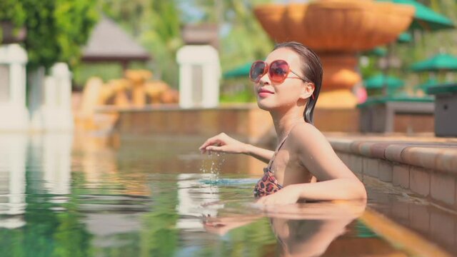 Pretty Asian model girl adjusting sunglasses inside swimming pool on blurred background shallow focus handheld in slow motion