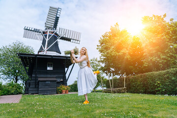 Woman dressed on traditional Dutch dress, wooden shoes yellow clogs klompen holding bouquet of Chamomile flowers with windmill on background. Retro vintage and countryside netherlands concept. 