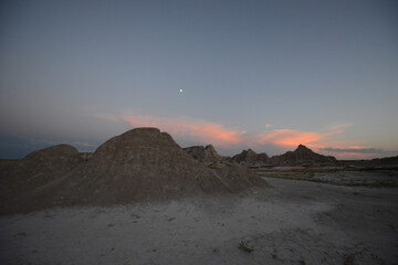 Landscape view of the unusual rocky terrain at Badlands National Park during twilight, with the moon in the sky