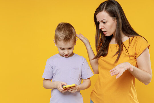 Sad young woman angry while child baby boy in violet t-shirt have fun hold use mobile cell phone play game. Mommy little kid son together isolated on yellow background studio Mother's Day love family.