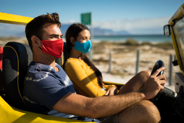 Happy caucasian couple wearing face masks sitting in beach buggy
