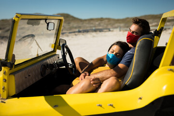 Happy caucasian couple wearing face masks sitting in beach buggy relaxing