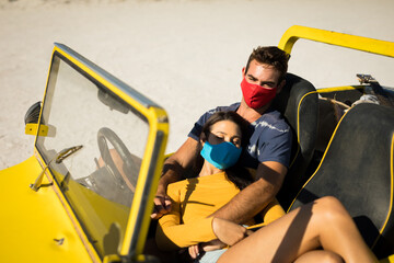 Happy caucasian couple wearing face masks sitting in beach buggy relaxing