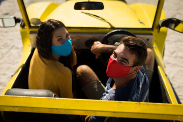 Happy caucasian couple wearing face masks sitting in beach buggy looking to camera