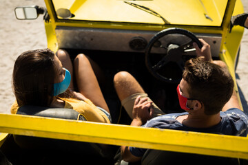 Happy caucasian couple wearing face masks sitting in beach buggy