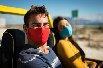 Happy caucasian couple wearing face masks sitting in beach buggy looking to camera