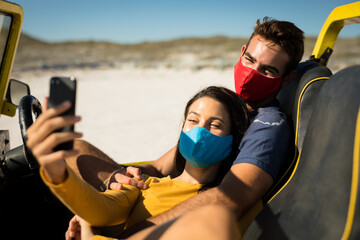 Happy caucasian couple wearing face masks sitting in beach buggy taking selfies