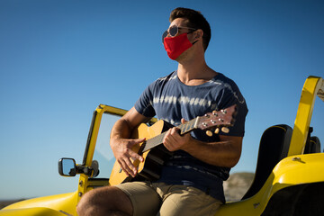Happy caucasian man wearing face mask sitting on beach buggy playing guitar