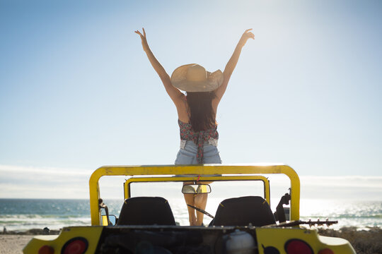 Caucasian woman sitting on beach buggy by the sea wearing straw hat looking toward sea with hands up - Powered by Adobe