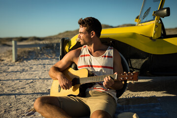 Happy caucasian man leaning against beach buggy by the sea playing guitar