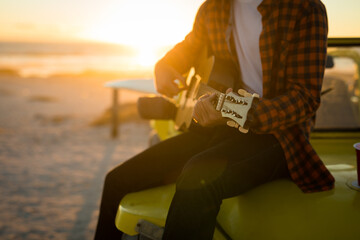 Midsection of caucasian man sitting on beach buggy by the sea playing guitar during sunset