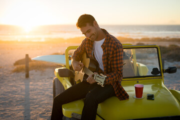 Happy caucasian man sitting on beach buggy by the sea playing guitar during sunset