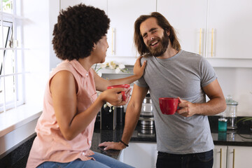 Happy diverse couple in kitchen drinking coffee and smiling