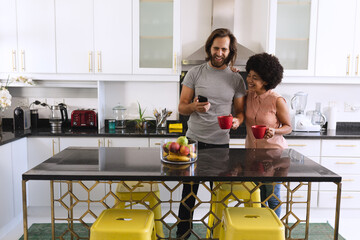 Happy diverse couple standing in kitchen drinking coffee and using smartphone