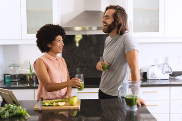 Happy diverse couple in kitchen smiling and drinking health drink