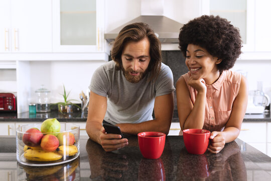Happy diverse couple standing in kitchen drinking coffee and using smartphone - Powered by Adobe