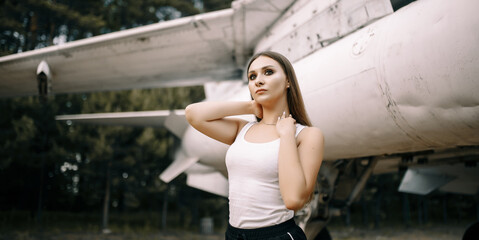beautiful young brunette girl stands on the background of old military aircraft. Girl in a white shirt and black pants in nature. Military equipment. Portrait half-length. girl posing