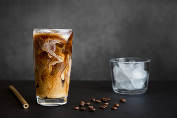 Iced coffee in tall glass with cream, container with ice, cocktail straw and coffee beans on dark background with copy space. Refreshing drink