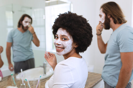 Portrait of happy diverse couple standing in bathroom wearing beauty masks