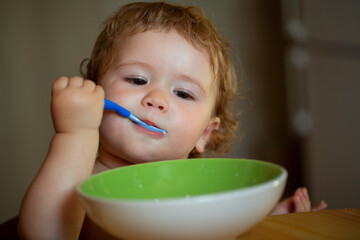 Portrait of funny little baby boy eating from plate holding spoon closeup. Baby holding a spoon while putting eat in his mouth.