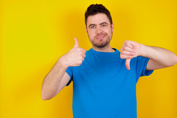 young handsome caucasian man wearing blue t-shirt against yellow background showing thumbs up and thumbs down, difficult choose concept - obrazy, fototapety, plakaty