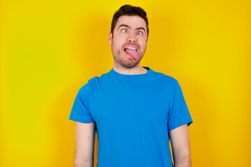 Obraz premium young handsome caucasian man wearing blue t-shirt against yellow background showing grimace face crossing eyes and showing tongue. Being funny and crazy