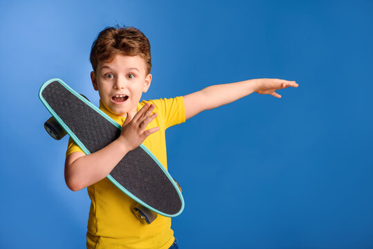 The kid boy in the yellow t-shirt is holding a penny board skateboard and laughing an pointing with hand on the blue background.