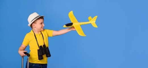 Happy little boy kid in a yellow t-shirt with a binocular is holding a yellow plane in one hand and a black suitcase in other on blue background