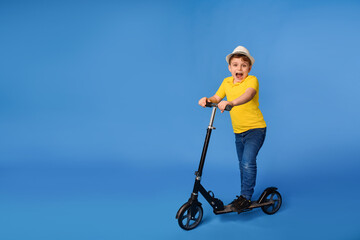 Little smiling boy in white hat and yellow t-shirt is raiding a kick scooter on a blue background.