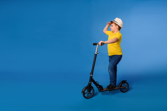 Little smiling boy in white hat and yellow t-shirt is raiding a kick scooter on a blue background.